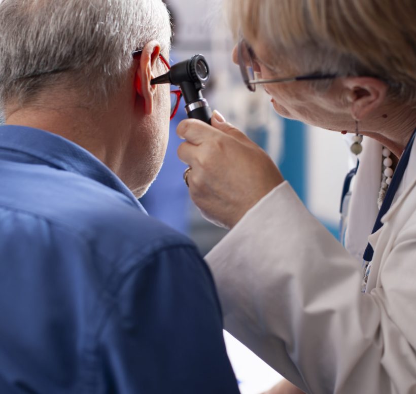 Experienced retired female physician inspecting ear of elderly male patient in clinic office. Closeup of skilled old doctor using otoscope during medical consultation of senior white man.